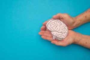 A brain shape made from paper in hands senior woman on a blue background. Awareness of Alzheimer's, Parkinson's disease, dementia, stroke, seizure, or mental health. Neurology and Psychology care