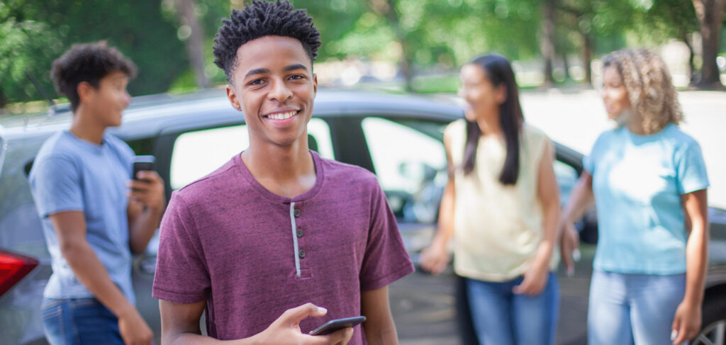 Portrait of young Black ethnicity teenager boy standing in front of car vehicle using smart phone with multiracial group of friends outside in parking lot