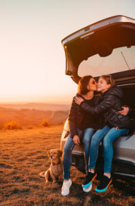 Mother kissing daughter while sitting in car trunk at hill and beside them is small brown dog