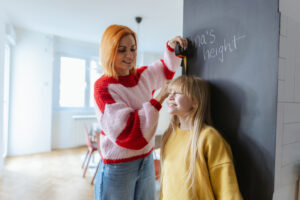 A loving mother helps her daughter measure height on a chalkboard in a sunny living room, capturing a warm, playful family moment of care and togetherness.