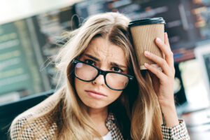 A tired young female programmer in glasses presses a takeaway coffee cup to her head, looking frustrated and exhausted while staring at code on multiple monitors in a busy office workspace.